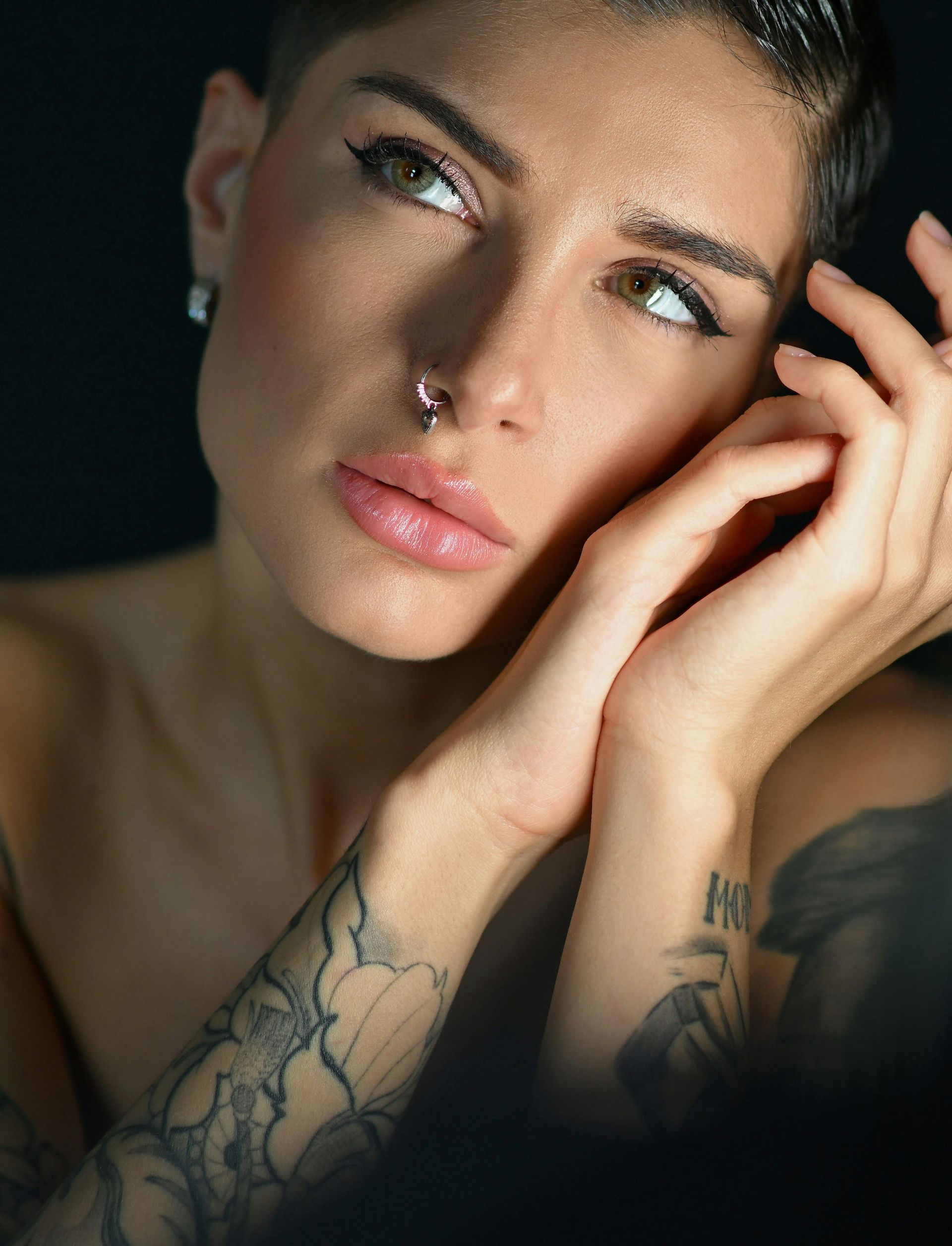Portrait of a tattooed person with a nose ring, resting their face on their hands in soft light
