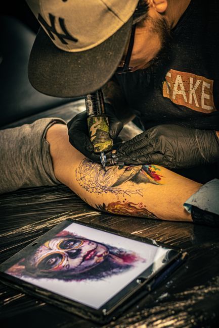 Tattoo artist in hat tattooing a client’s arm beside a framed portrait reference