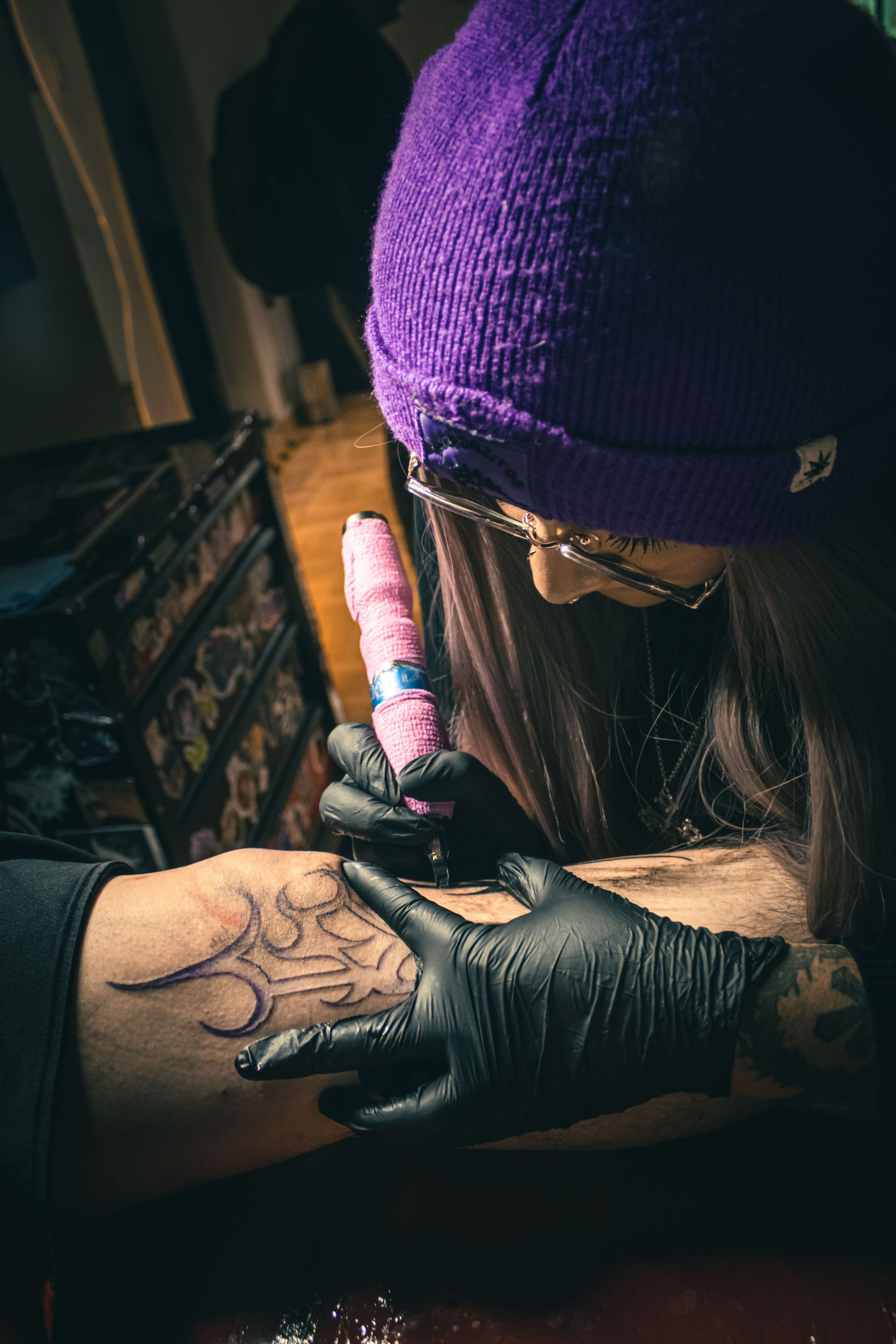 Tattoo artist in purple beanie tattooing a hand with a black glove