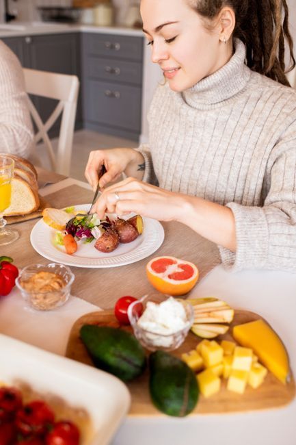 Person plating food at a dining table with fresh fruit and vegetables in a bright kitchen