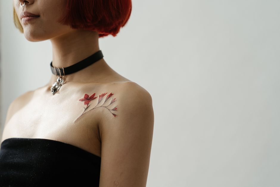Woman in black strapless top wearing a choker, with a red tattoo on her shoulder against a plain background