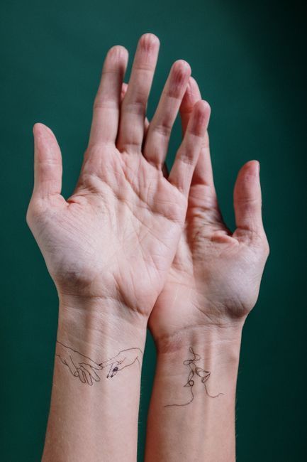 Two raised hands with slender fingers against a teal background, wrists showing fine line tattoos