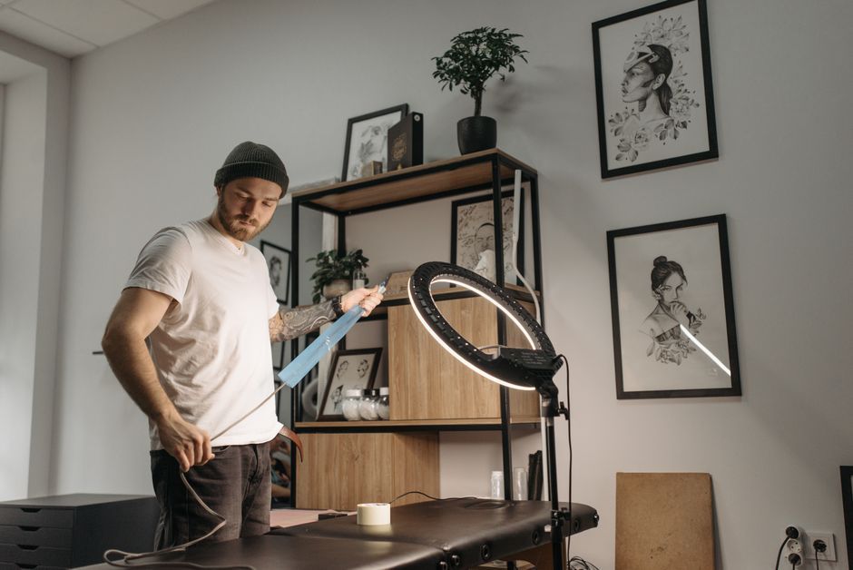 Man standing by a treatment table in a bright studio, holding a phone near a ring light and shelving unit.