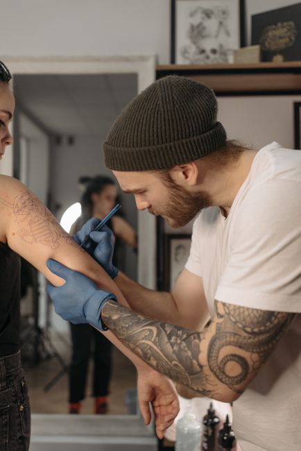 Tattoo artist wearing gloves works on a client’s arm in a studio, with a mirror visible in the background.