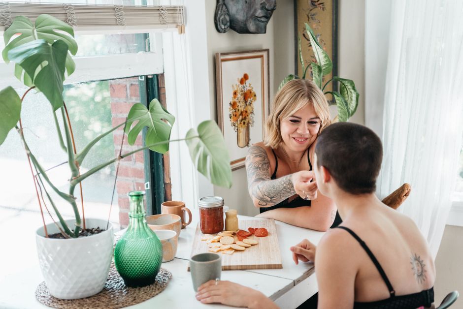 Woman applies makeup at a vanity near a window, with plants, mirrors, and cups on the table.