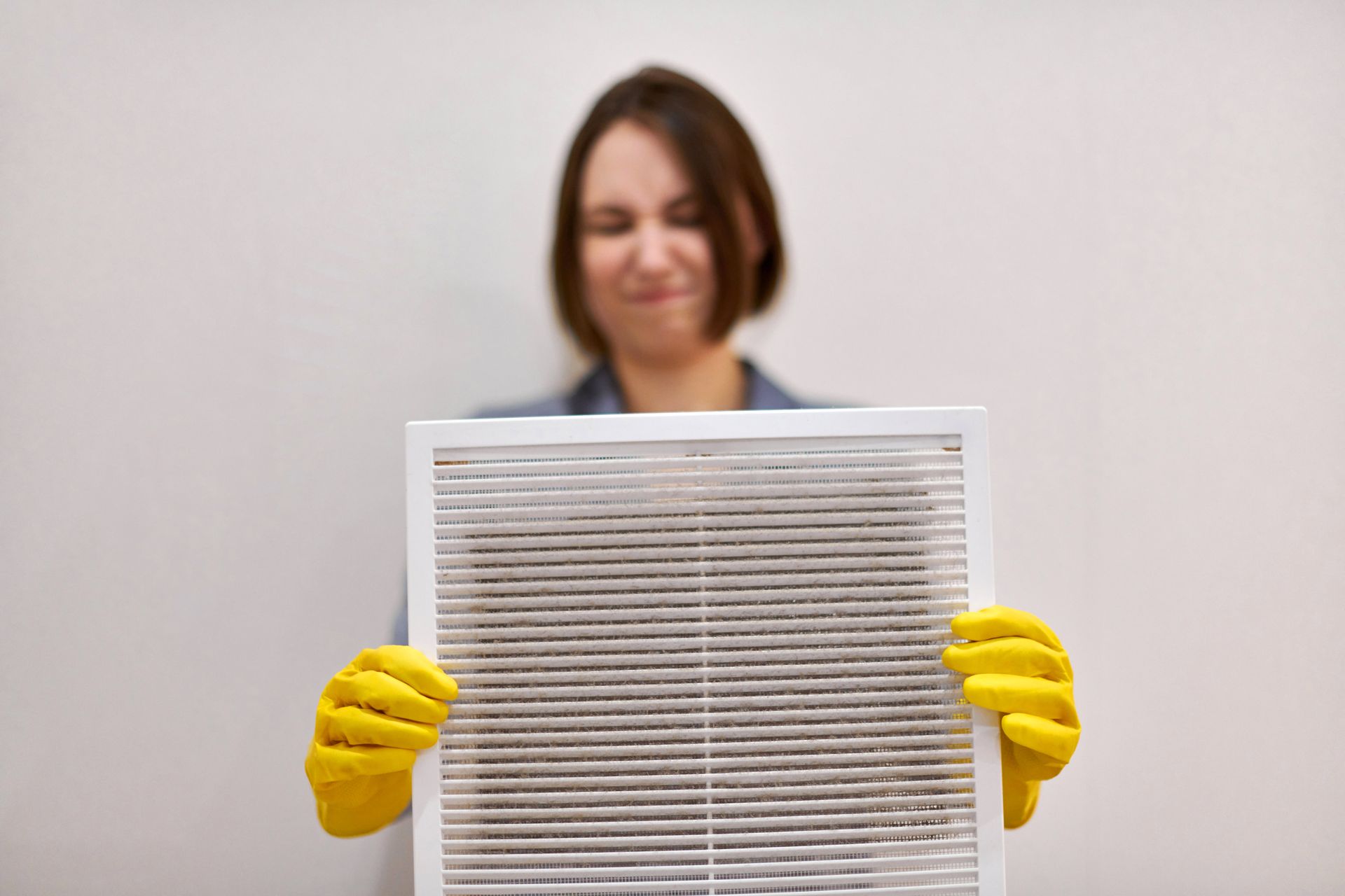 Woman in yellow gloves holds a dirty air filter, frowning. White background.