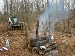 A fire in the woods with a tractor in the background.
