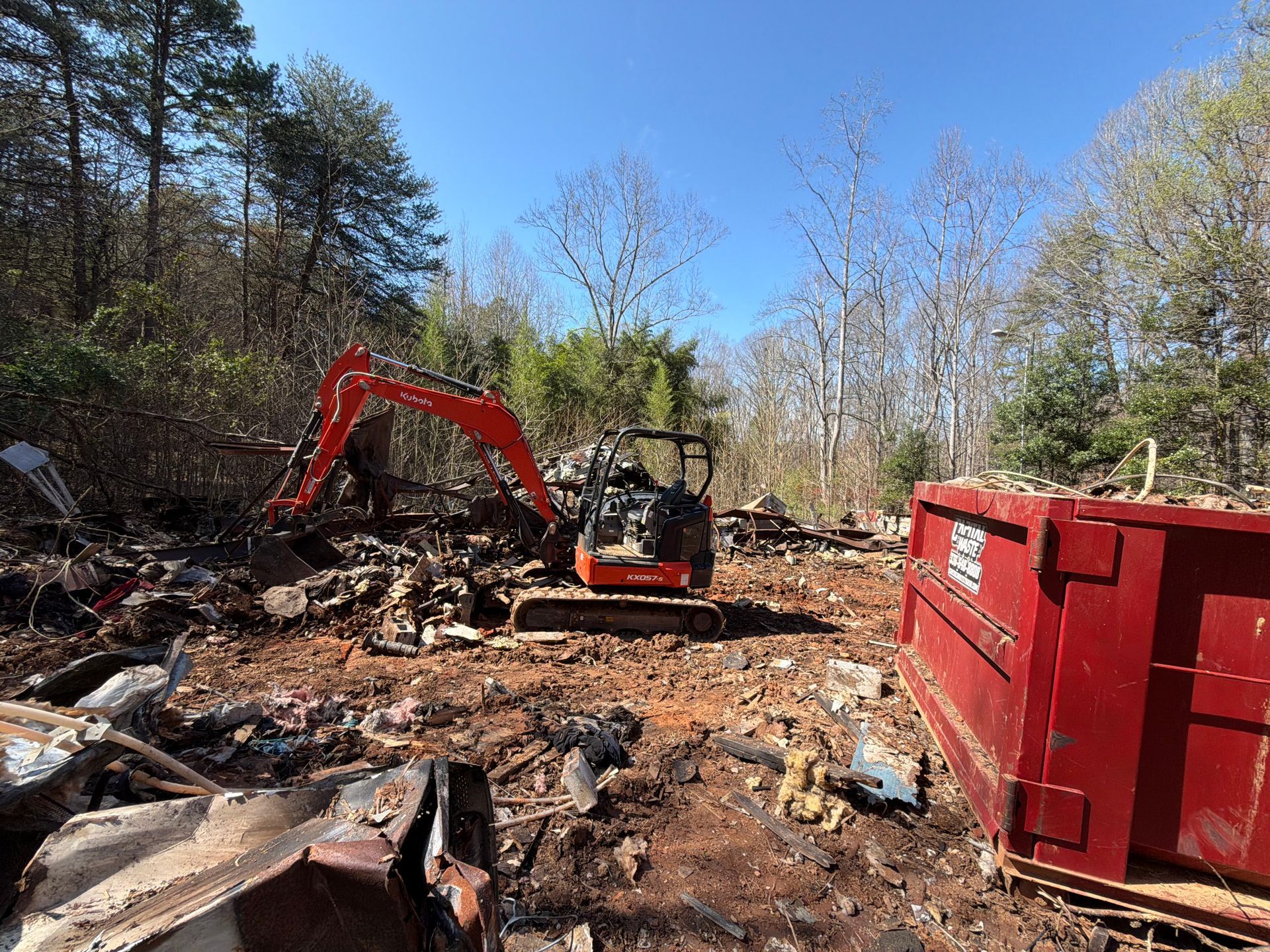 Orange excavator clearing debris in a wooded lot beside a large red dumpster under a blue sky