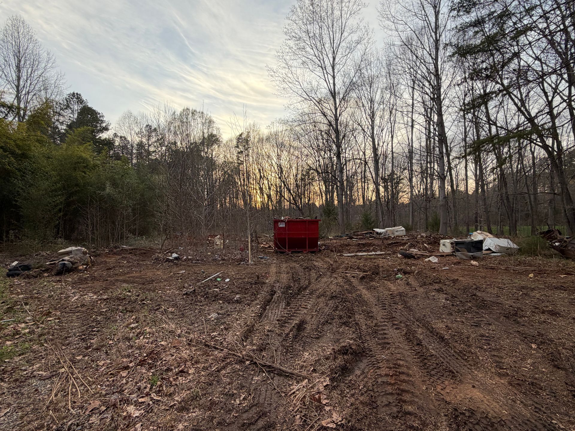 Red dumpster sits in a muddy clearing with bare trees at dusk.