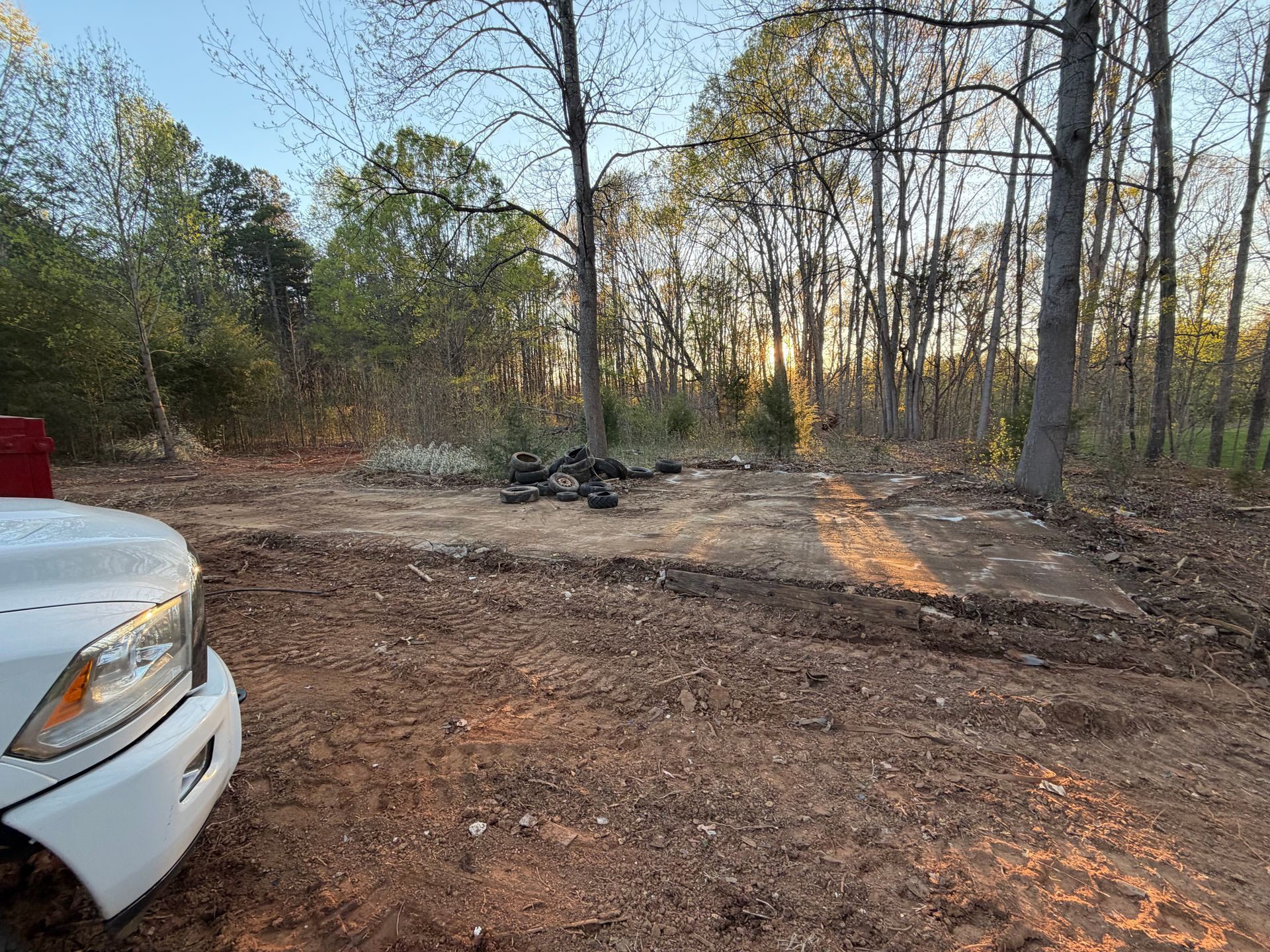 White vehicle at the edge of a wooded dirt clearing with a small fire pit and low sun through bare trees.