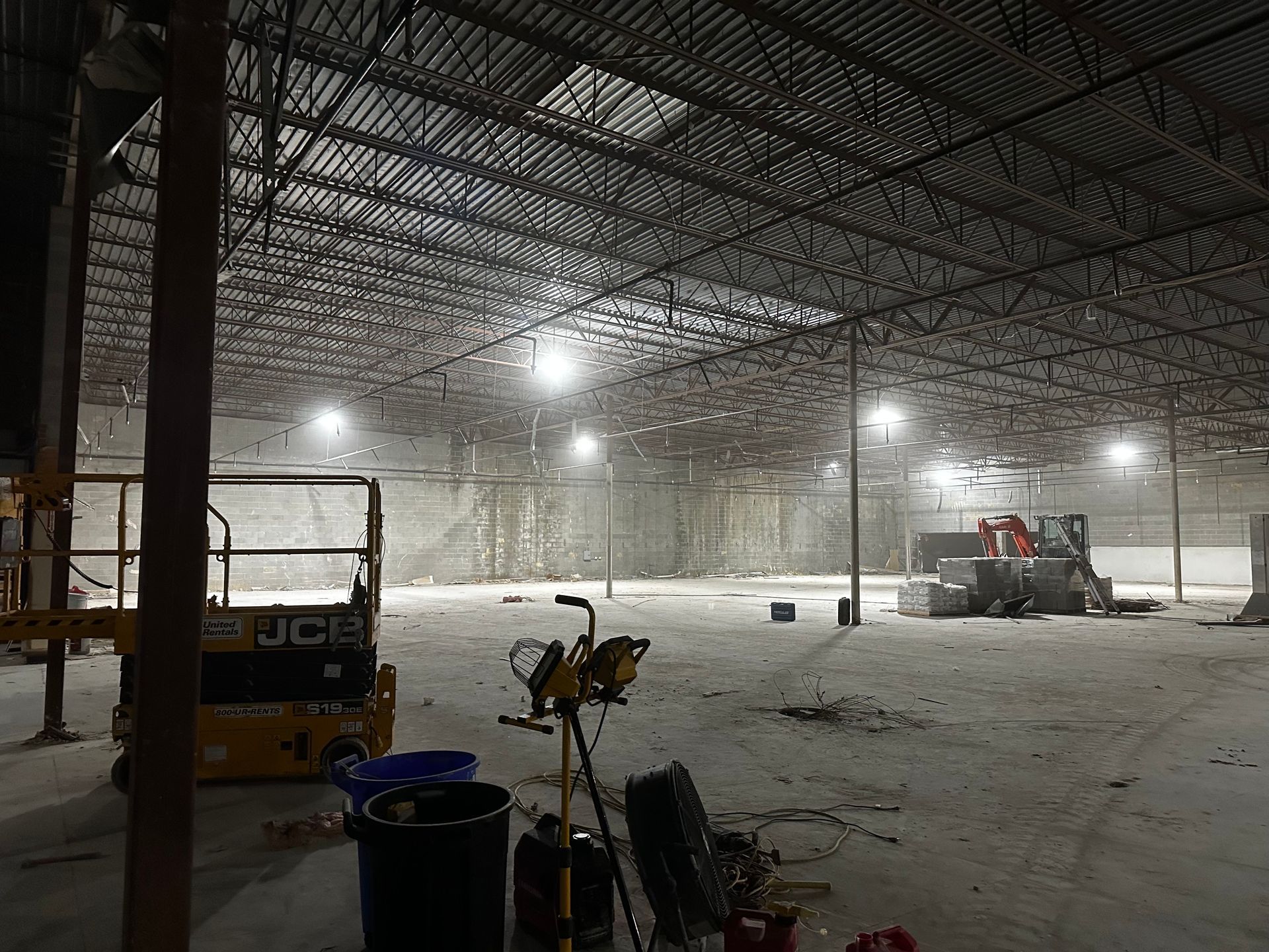 Construction site: large open warehouse interior with equipment, bright overhead lights, and exposed ceiling structure.