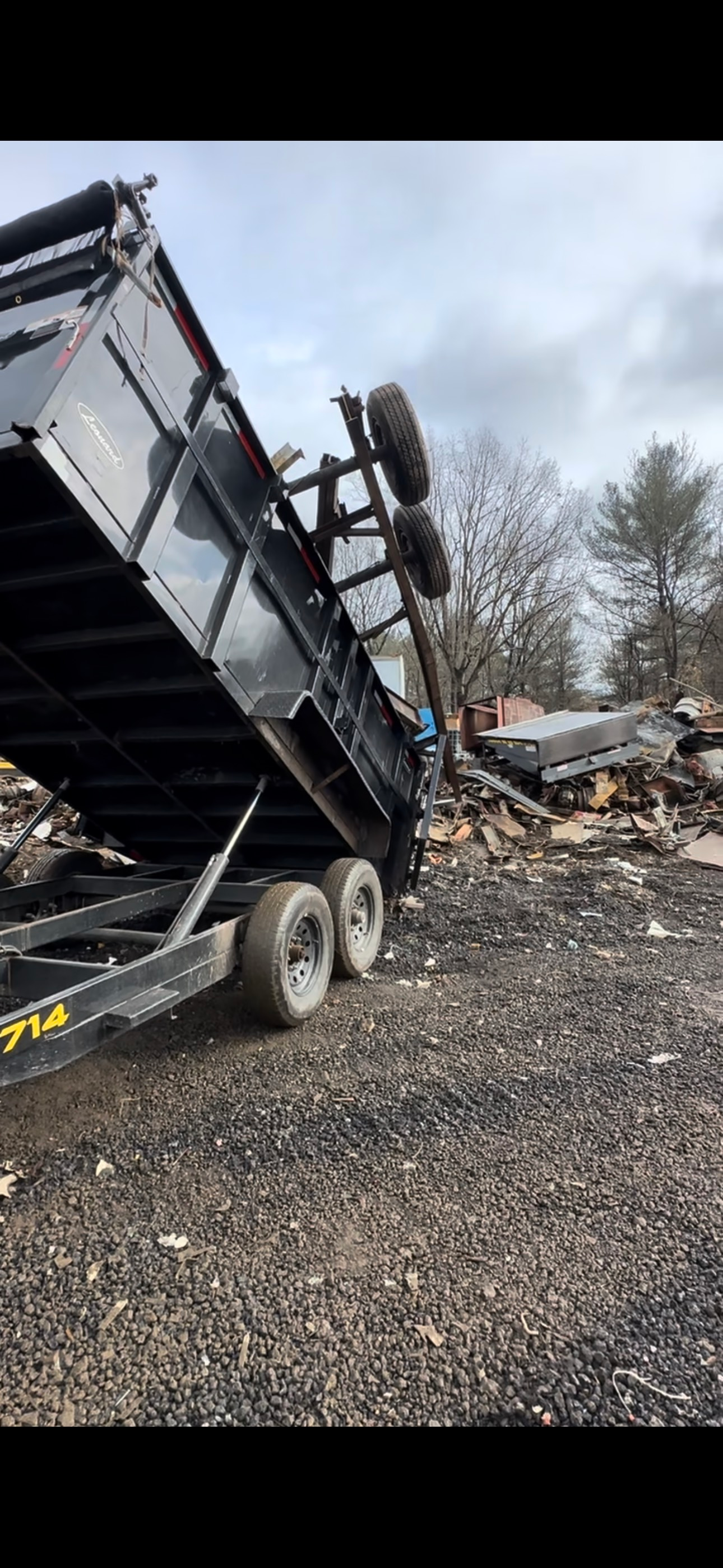 A black dump trailer unloading debris onto a gravel surface. Trees are visible in the background.