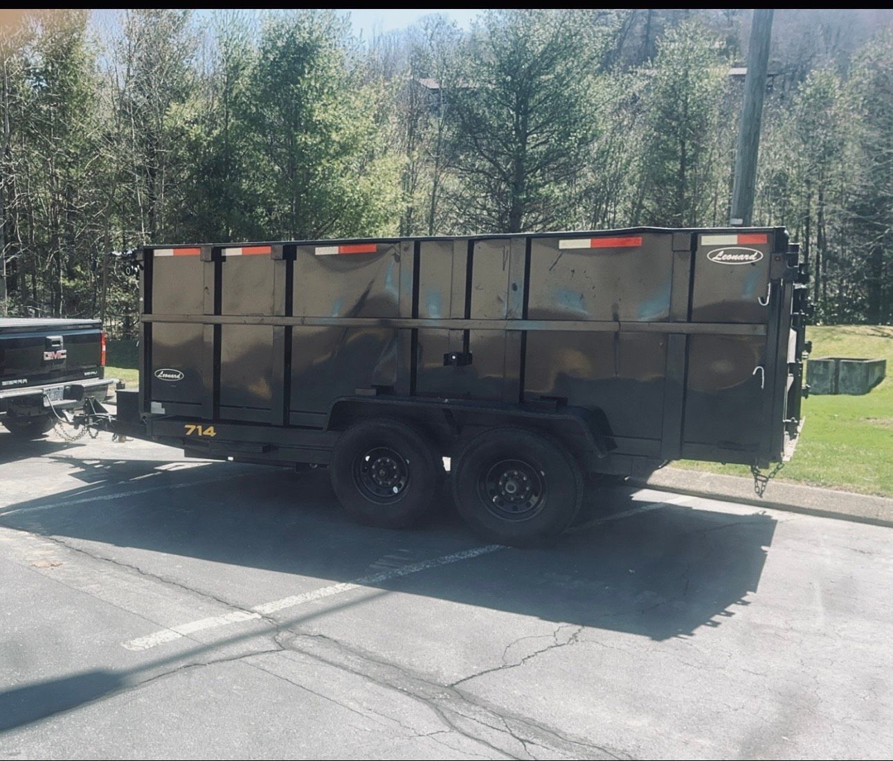 Black dumpster trailer parked in a paved lot, with trees in the background.
