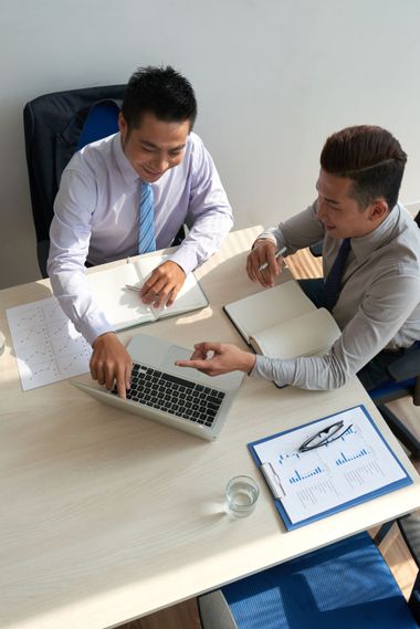 Two colleagues in business attire sit at a table, looking at a laptop and documents while collaborating in an office.
