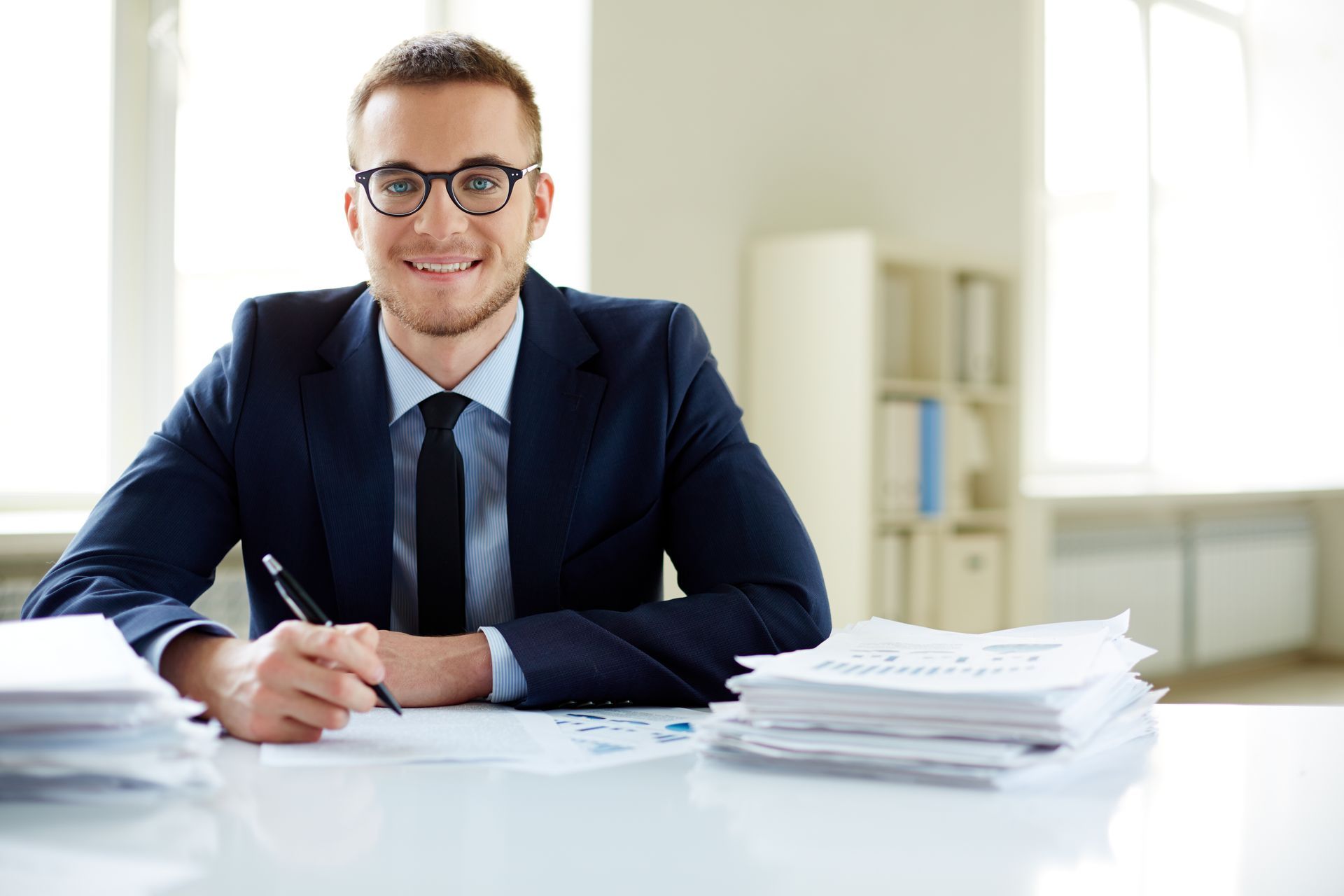 A smiling professional in a suit and glasses sitting at a desk with stacks of documents, holding a pen.