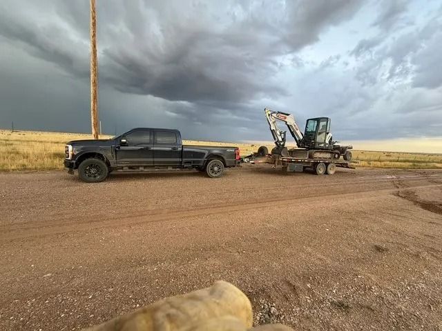 Black pickup with trailer and excavator on a dirt road under a cloudy sky in open fields