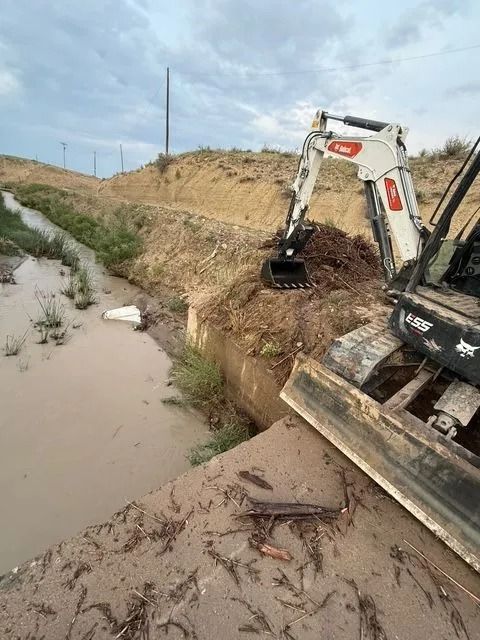 Excavator digging a muddy canal bank beside a water-filled ditch under a cloudy sky