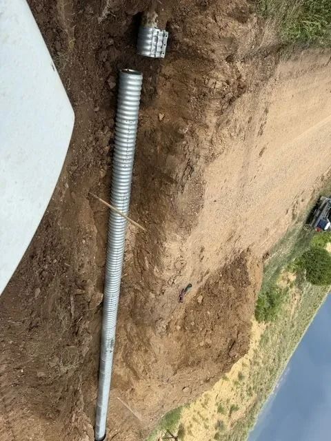 Long metal pipe laid in a dirt trench beside a concrete wall near a road and grassy slope.