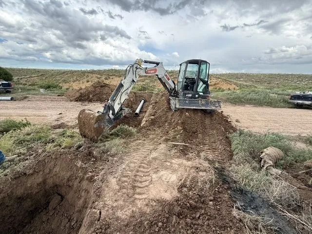 Excavator digging a dirt trench beside a muddy road under a cloudy sky