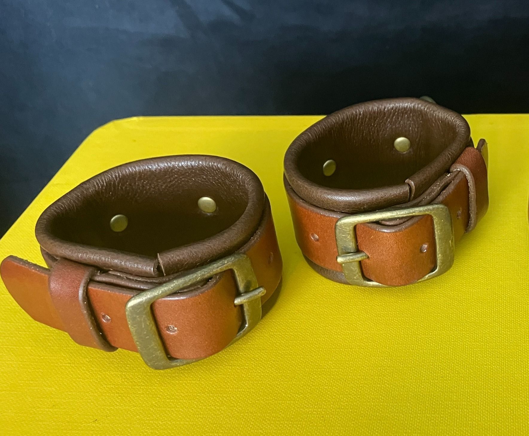 Two brown leather wrist cuffs with brass buckles, resting on a yellow surface.