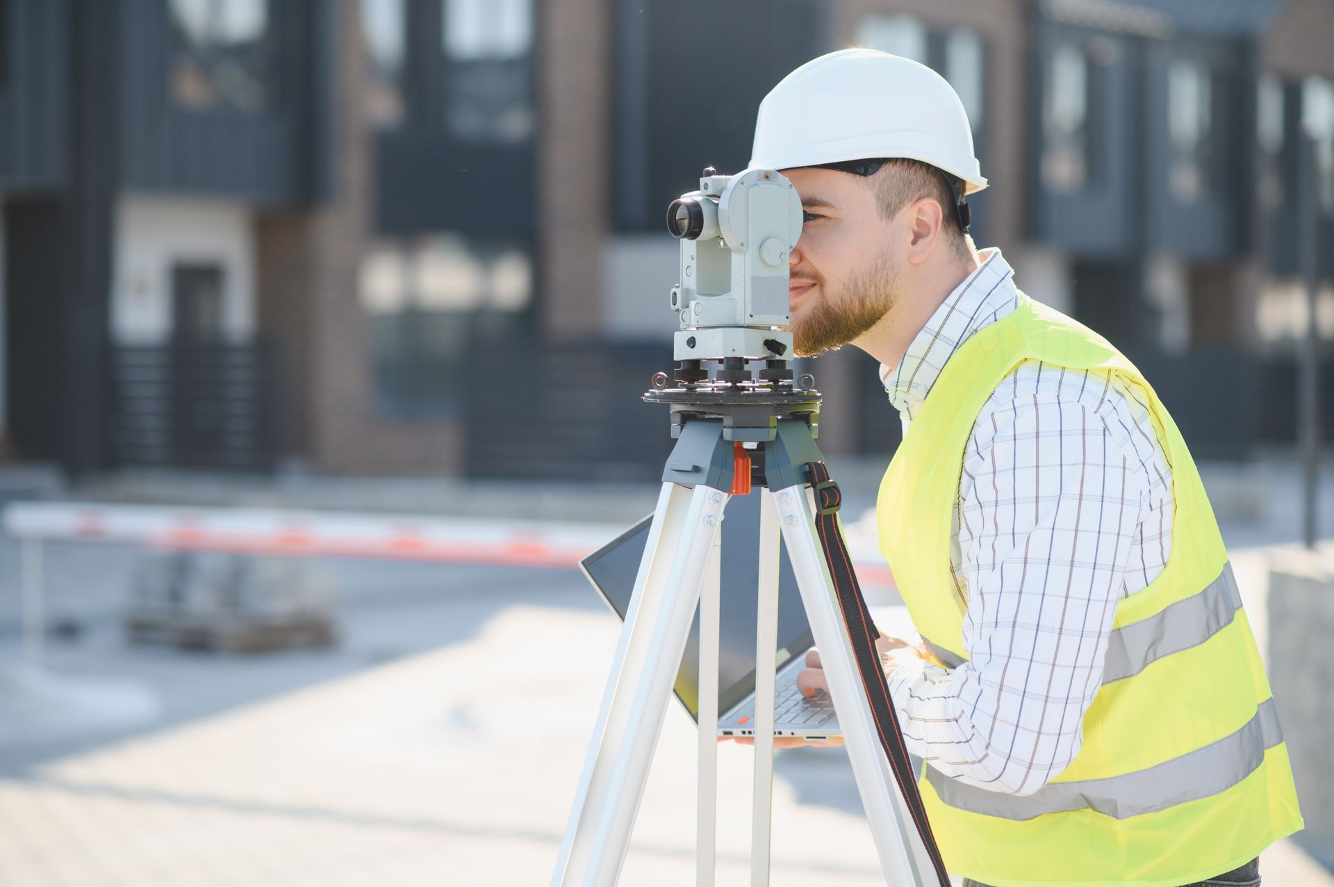 Surveyor in hard hat and safety vest using a tripod-mounted measuring instrument at a construction site