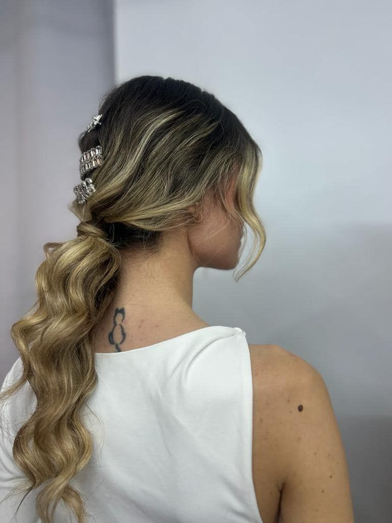 Donna con capelli biondi ondulati raccolti in una coda di cavallo laterale, ornati da forcine decorative. Top bianco su sfondo bianco.