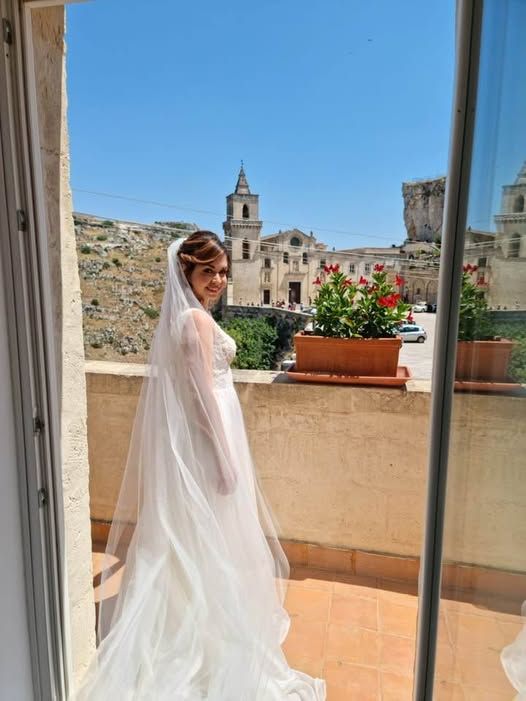 Sposa in abito bianco su un balcone, con una chiesa e una città sullo sfondo sotto un cielo azzurro.