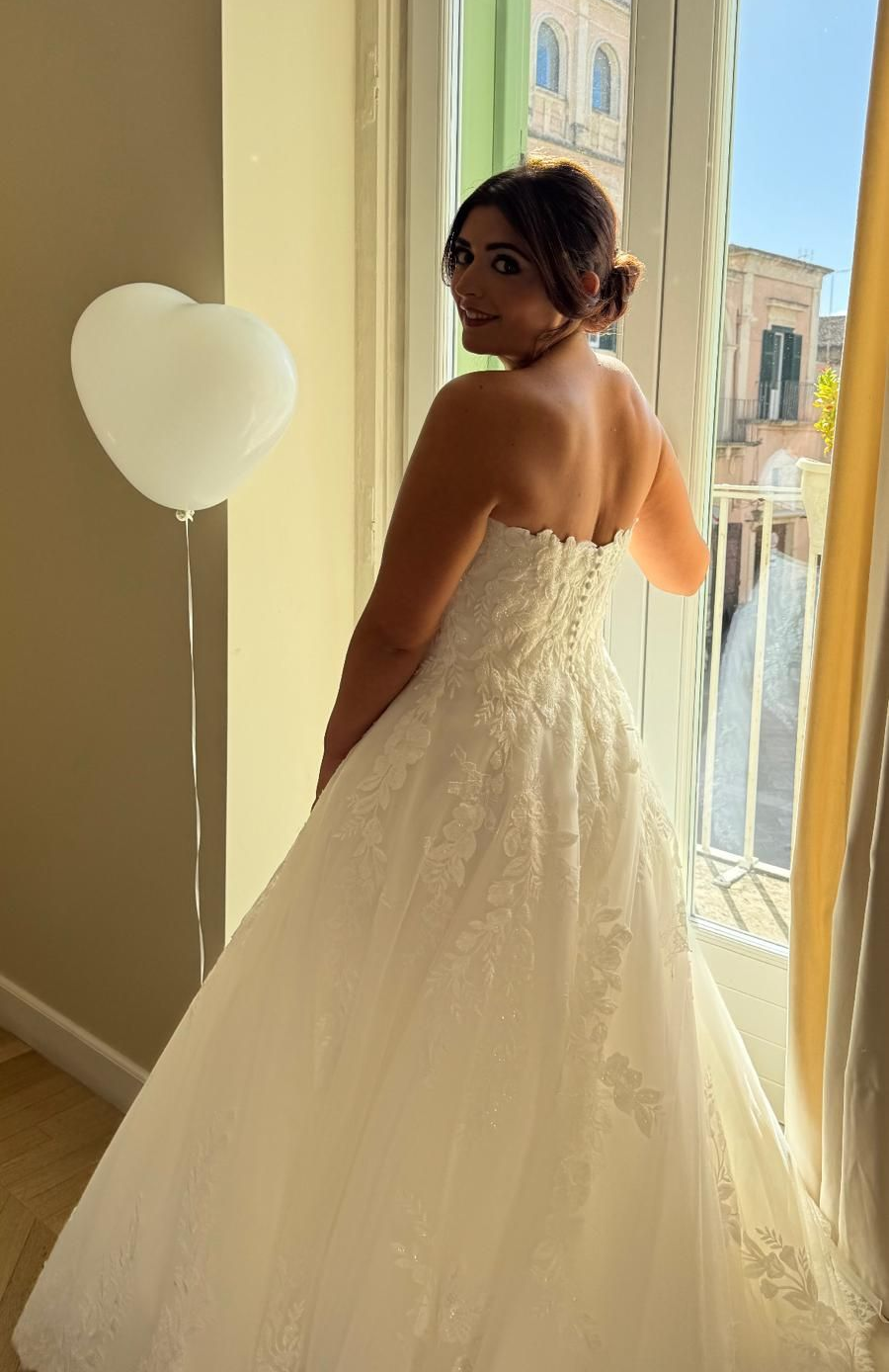 Donna in abito da sposa bianco senza spalline, girata verso l'osservatore da una finestra, con un palloncino a forma di cuore.