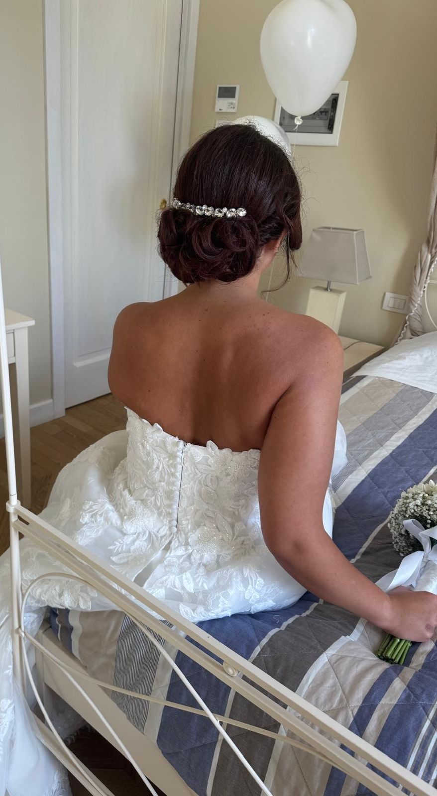 Sposa in abito bianco con dettagli elaborati, seduta su un letto. Acconciatura raccolta con fermaglio per capelli.