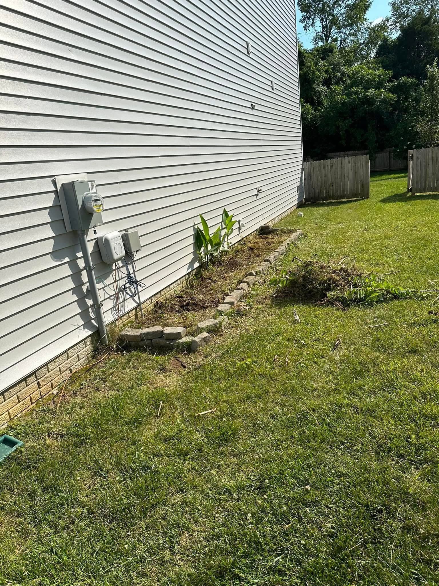 Side of a white house with utility meters, a narrow planting bed, and a grassy yard in sunlight