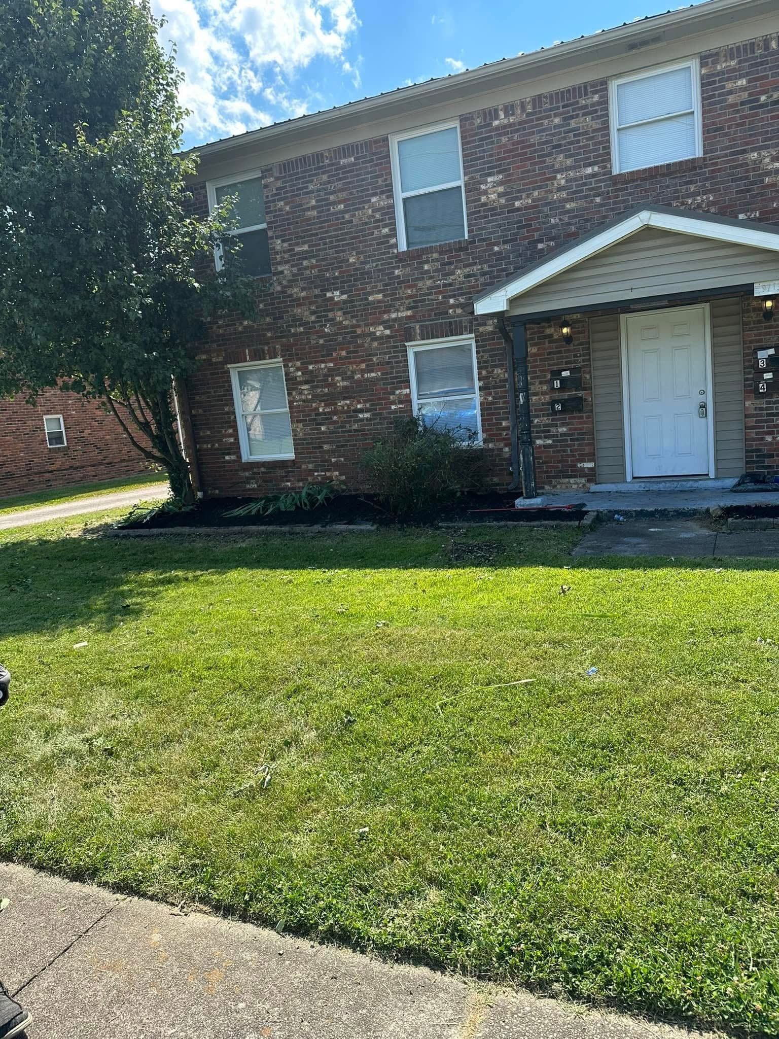 Two-story brick apartment building with white doors and windows, front lawn, and sidewalk in bright sunlight.