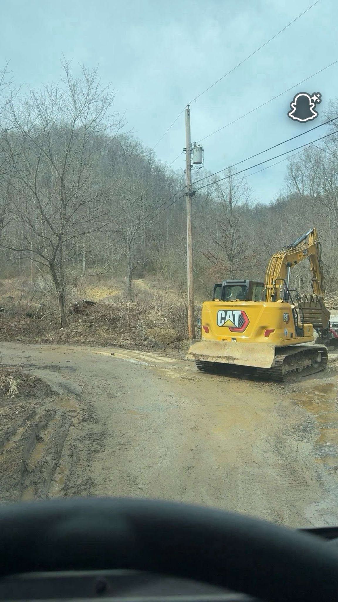 Yellow excavator on a muddy roadside near bare trees and utility poles, viewed from inside a vehicle