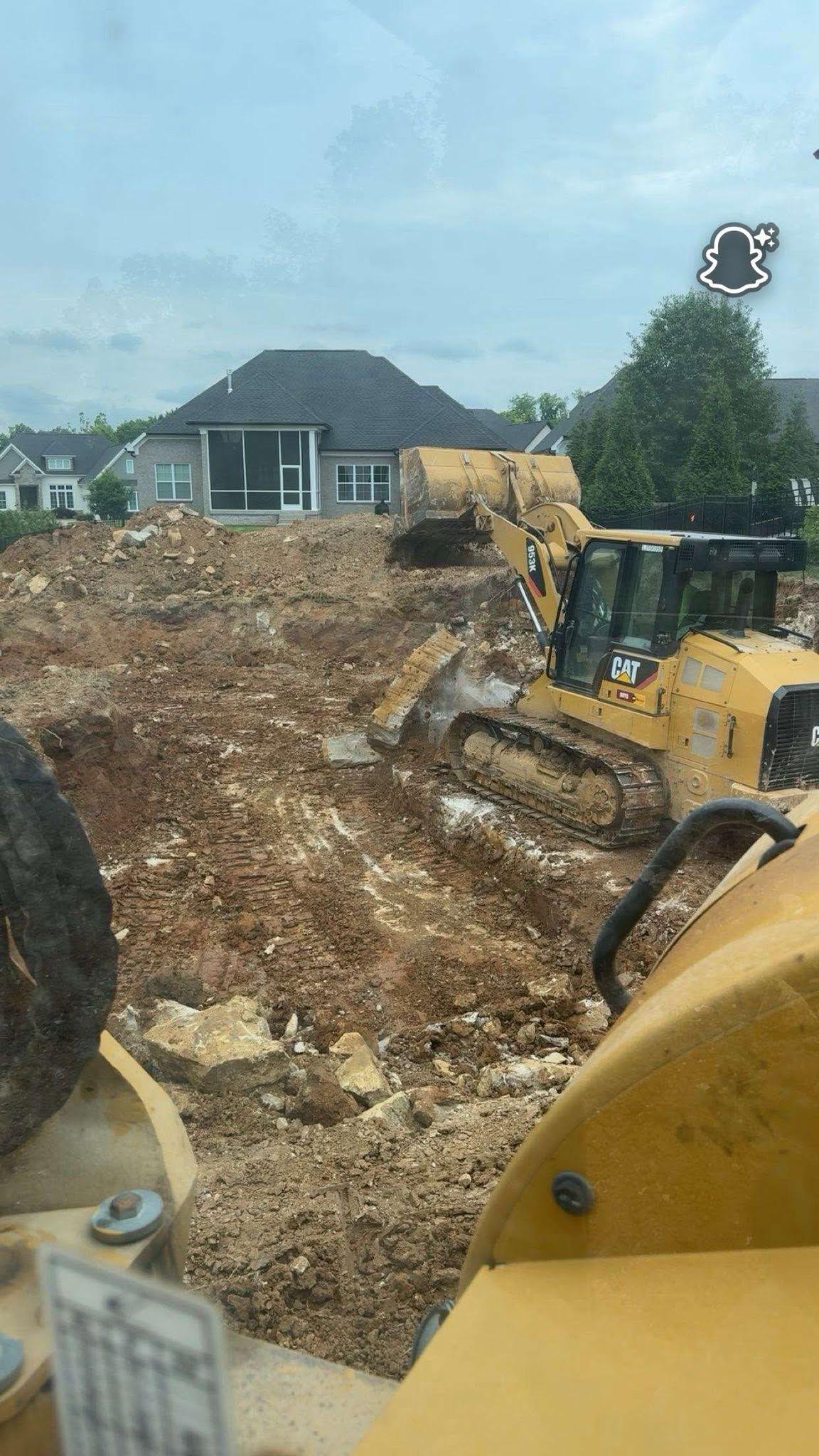 Construction site with excavator clearing dirt near houses under a cloudy sky