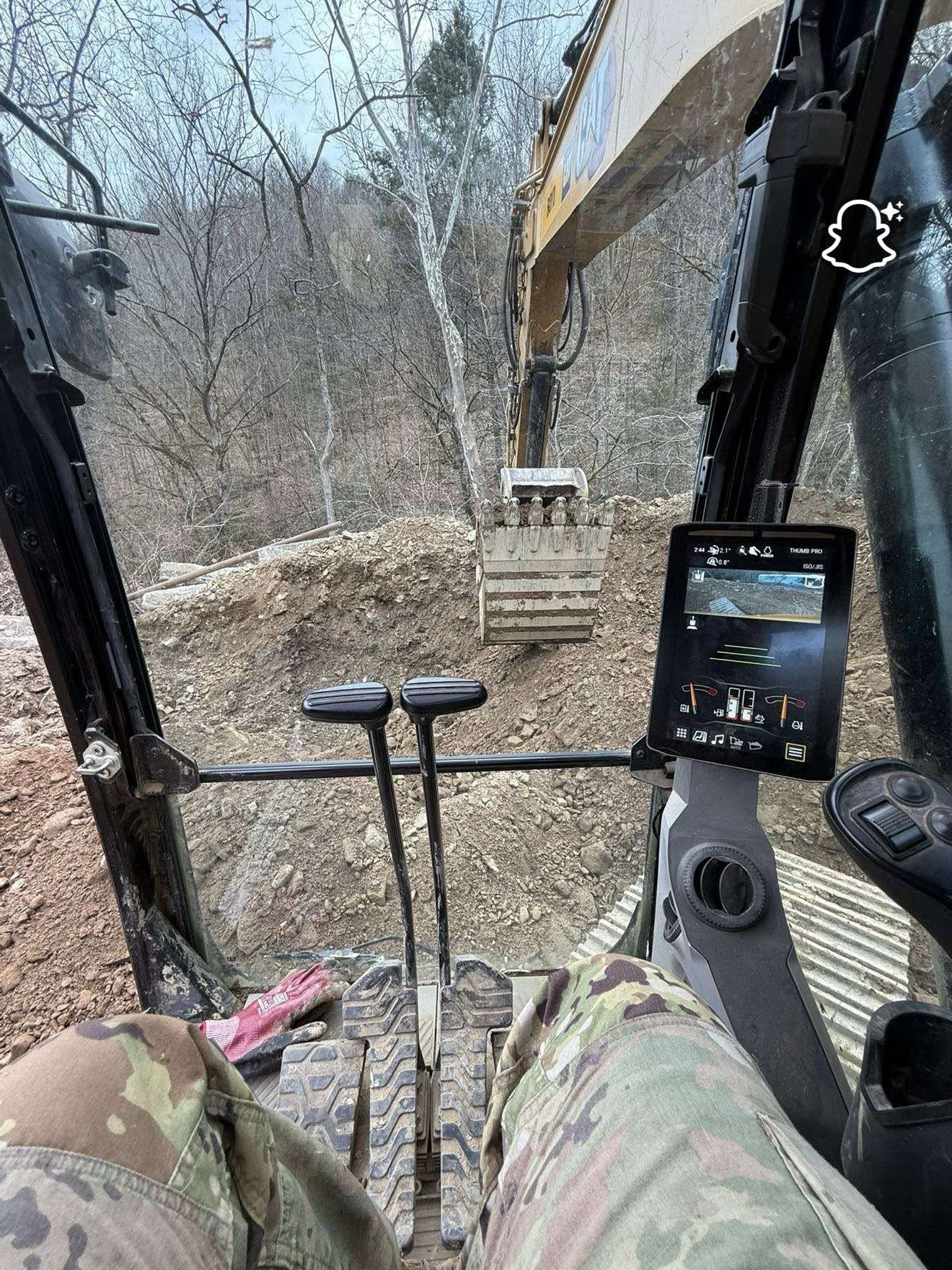 Inside a excavator cab, looking at the boom over a muddy wooded work site.