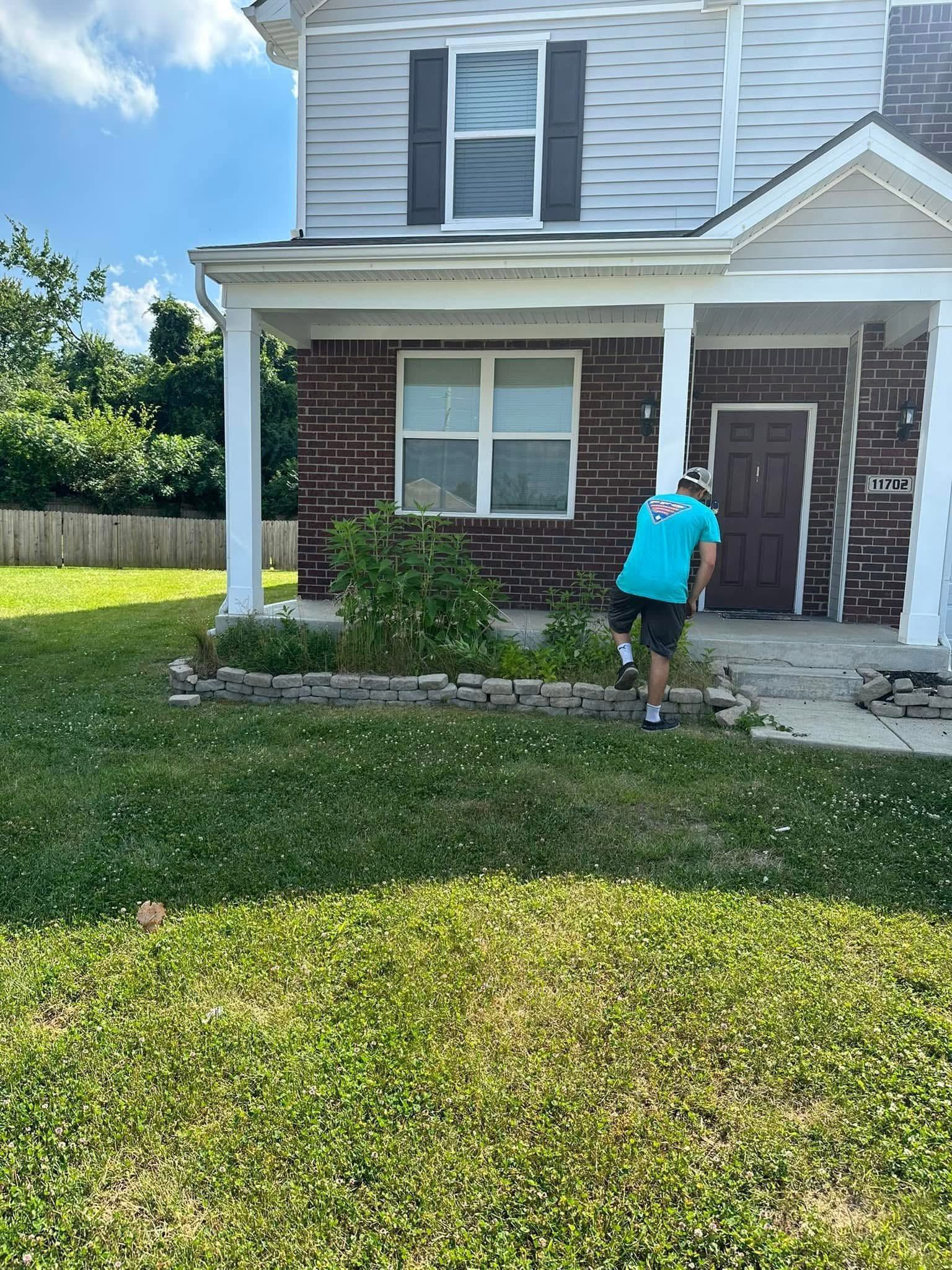 Person trimming shrubs in front of a brick house with a green lawn and porch