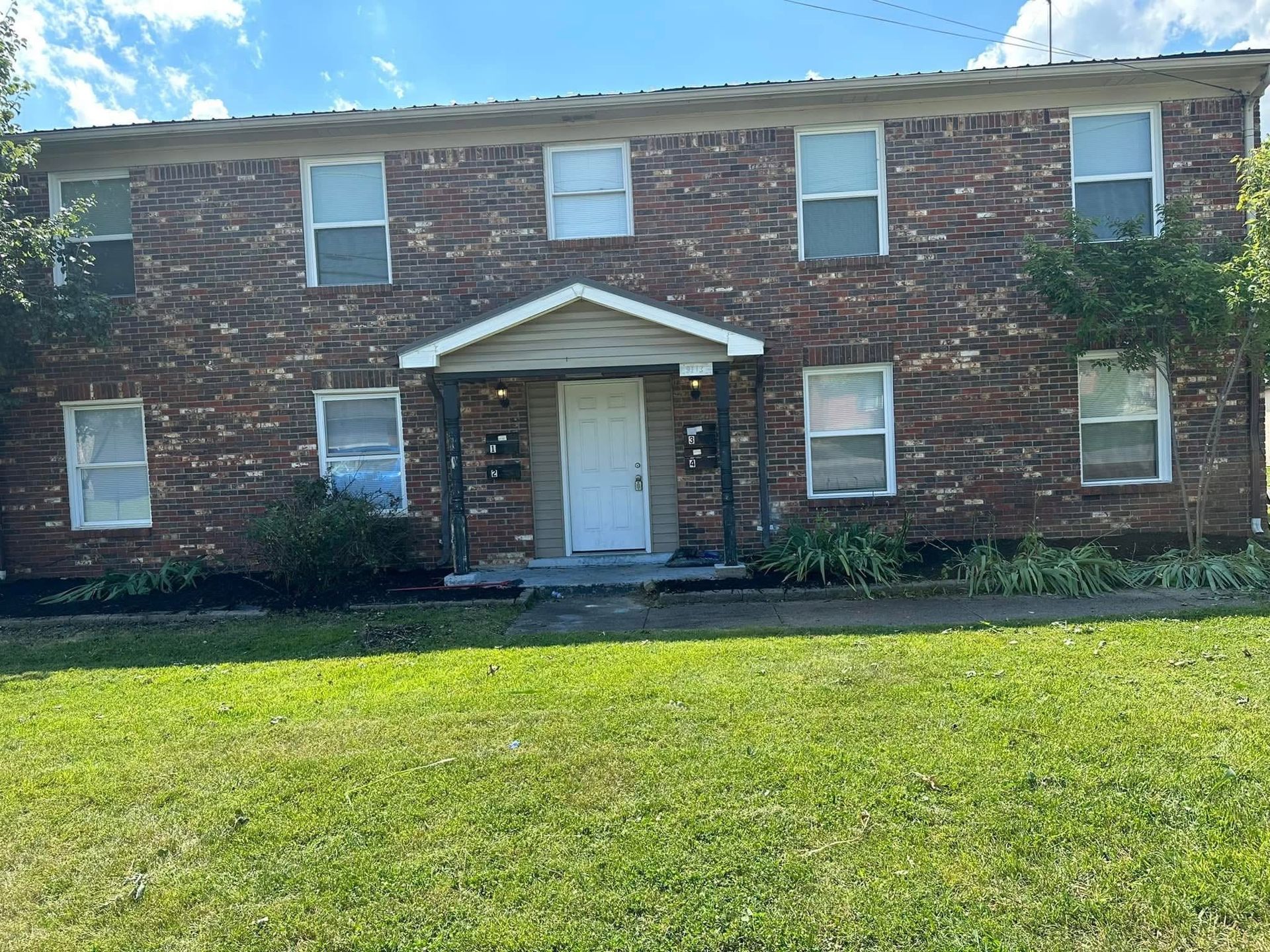 Brick two-story building with a white front door, steps, and green lawn under a blue sky
