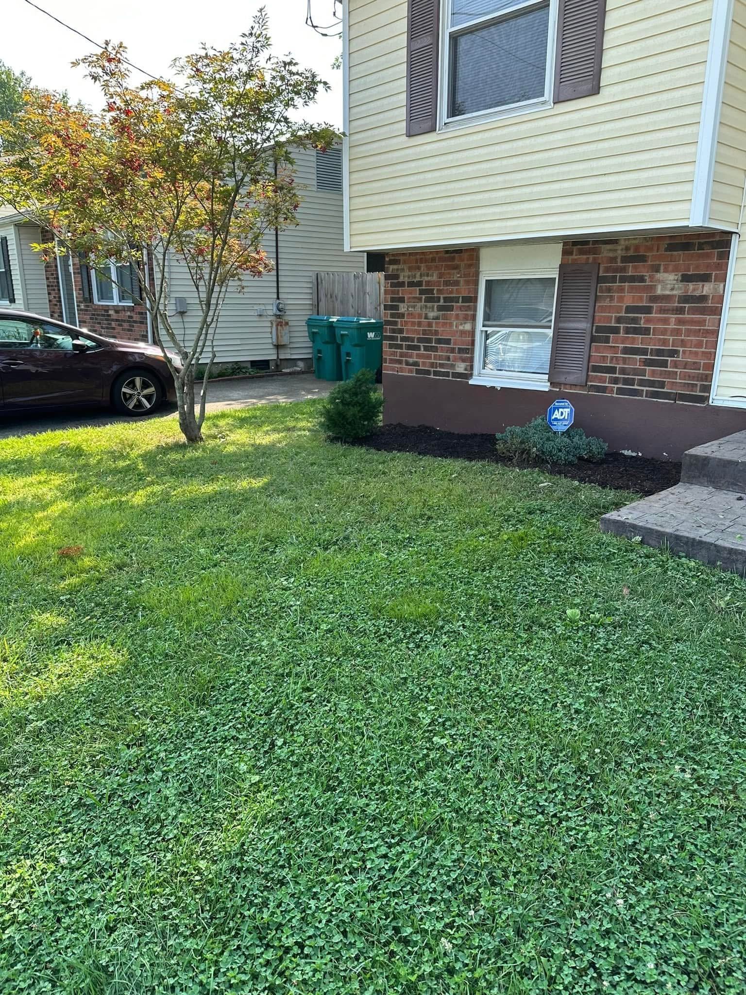 Front yard of a house with green grass, a tree, and a parked black car beside brick siding.