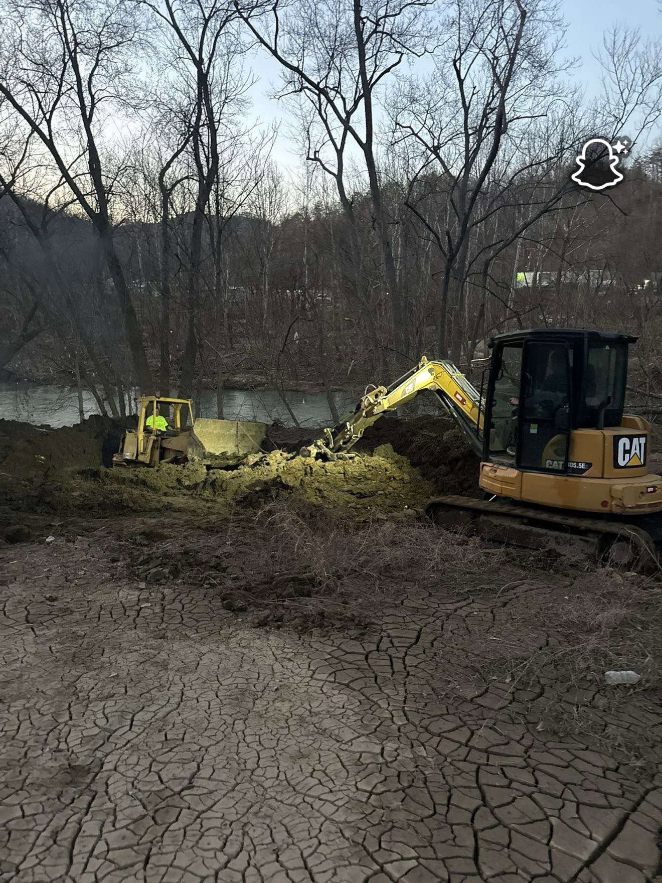 Two excavators dig in a muddy, leafless wooded area beside cracked ground.