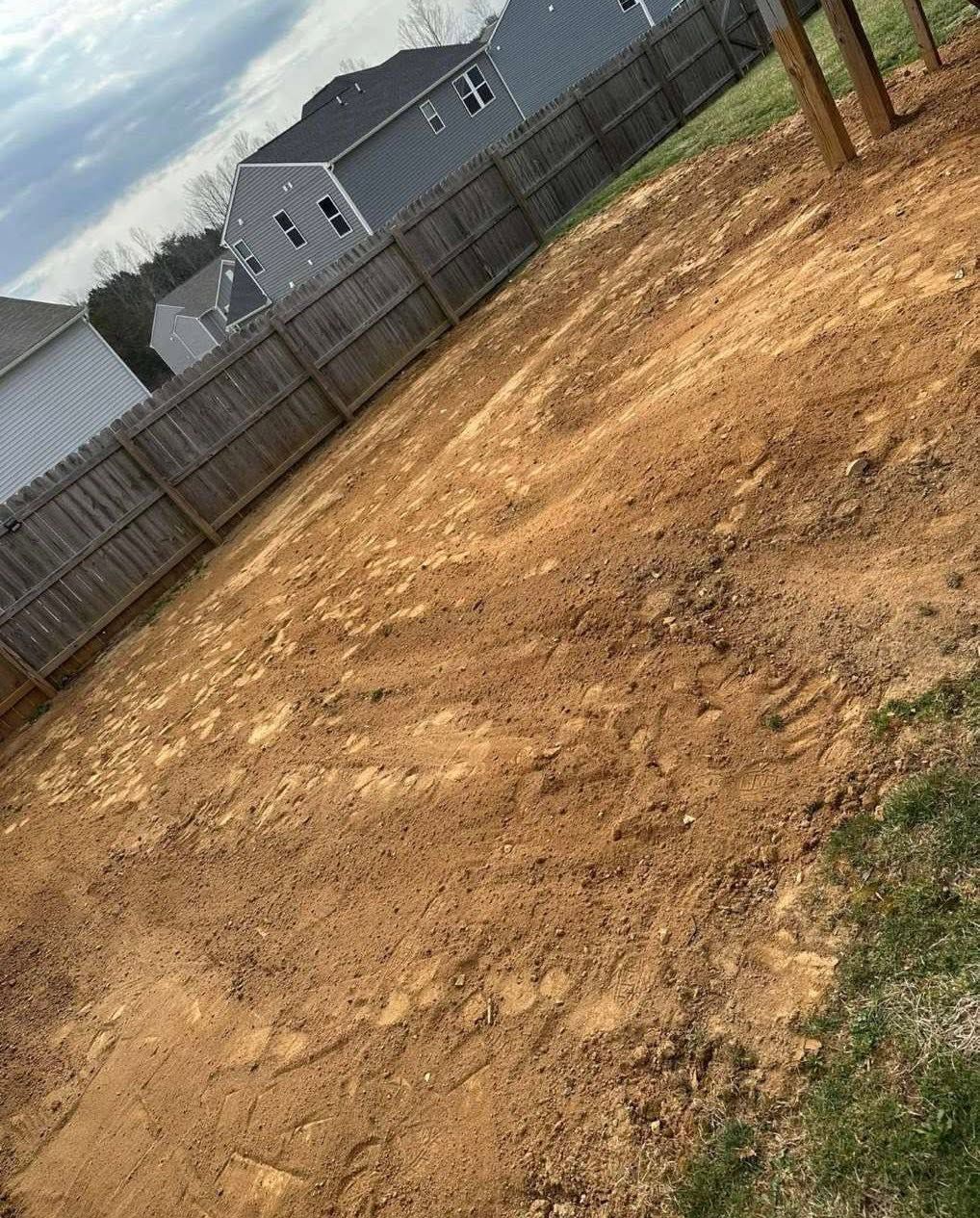 Bare dirt backyard beside a wooden fence and a gray house under a cloudy sky