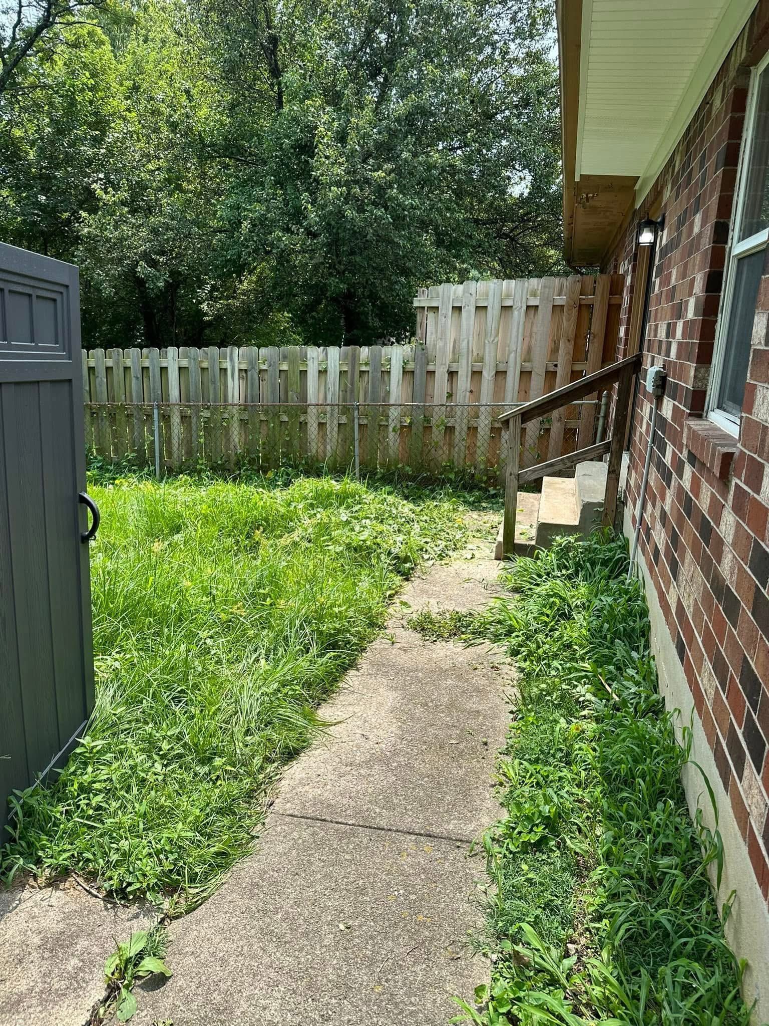 Narrow concrete path beside brick house leading to a wooden fence and grassy backyard.