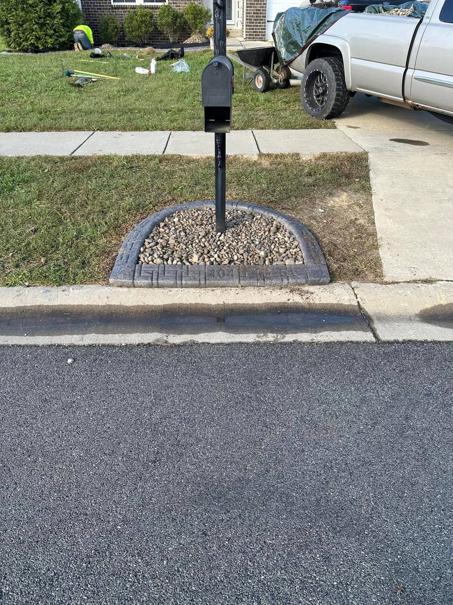 Mailbox on a small rock bed beside a sidewalk and curb, with a parked pickup truck in the driveway.