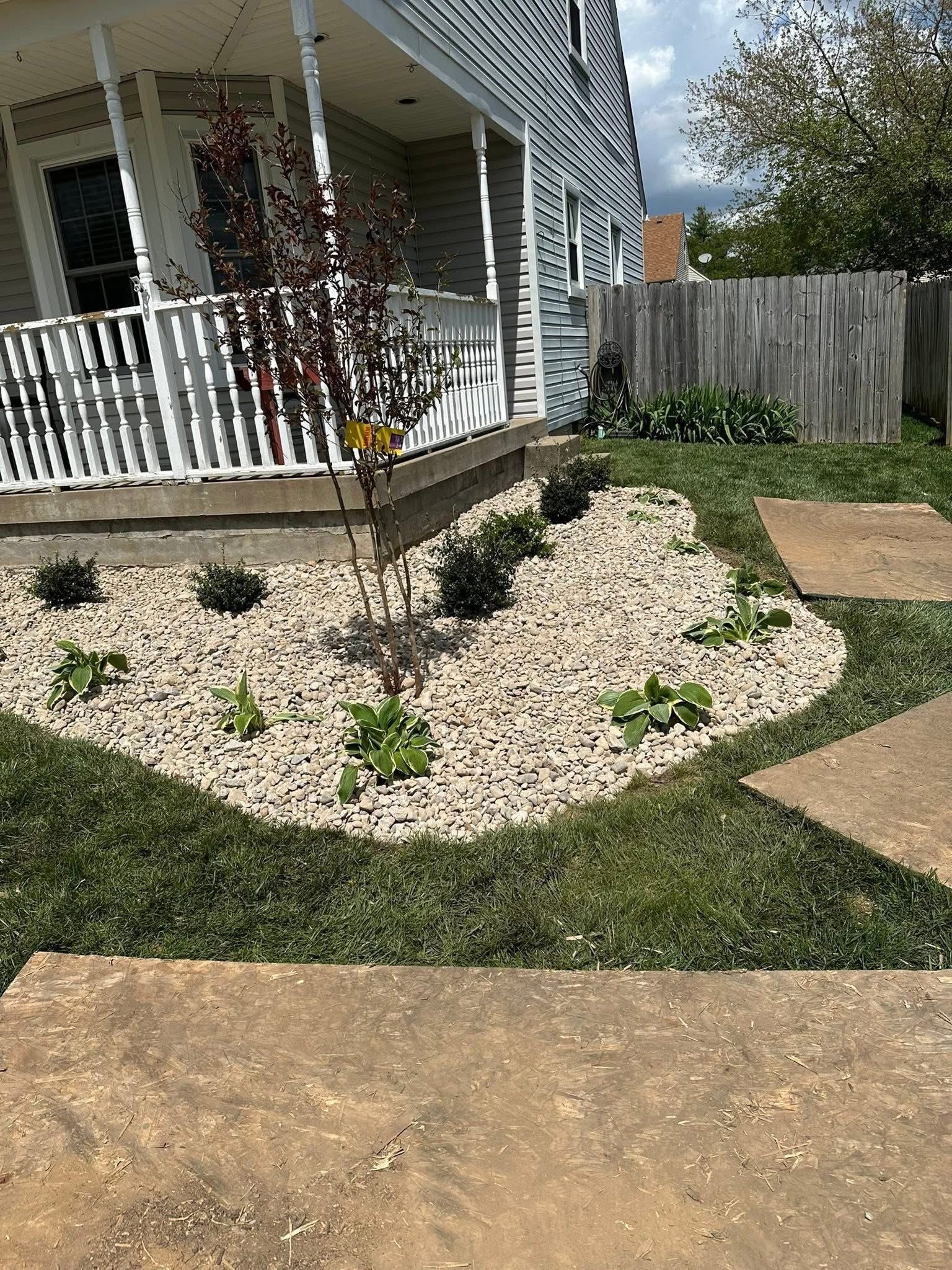 Front yard landscaping with white stone bed, small shrubs, a young tree, and a porch in the background
