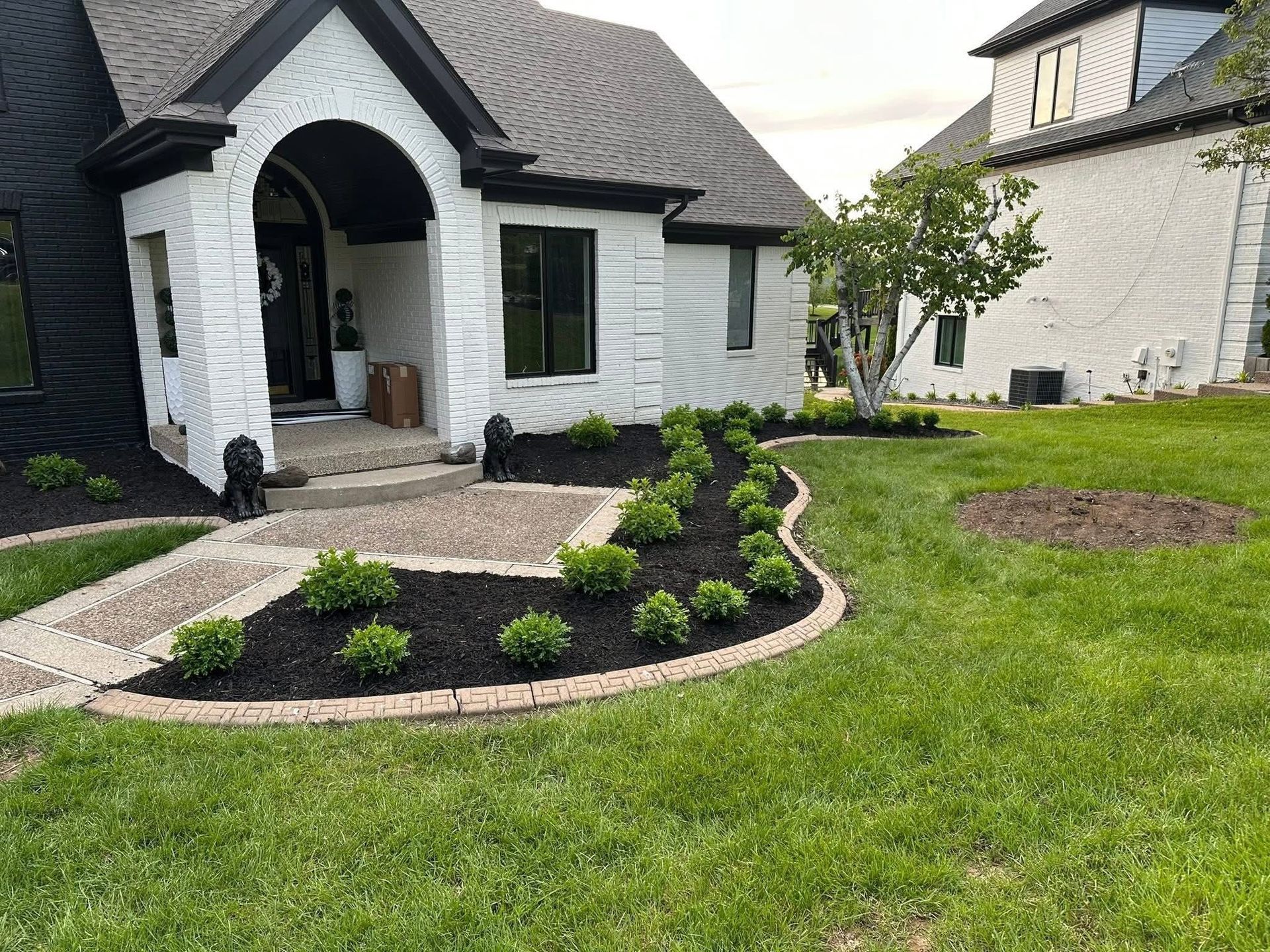 Front yard landscaping with curved garden beds and a white house with black trim and arched porch.