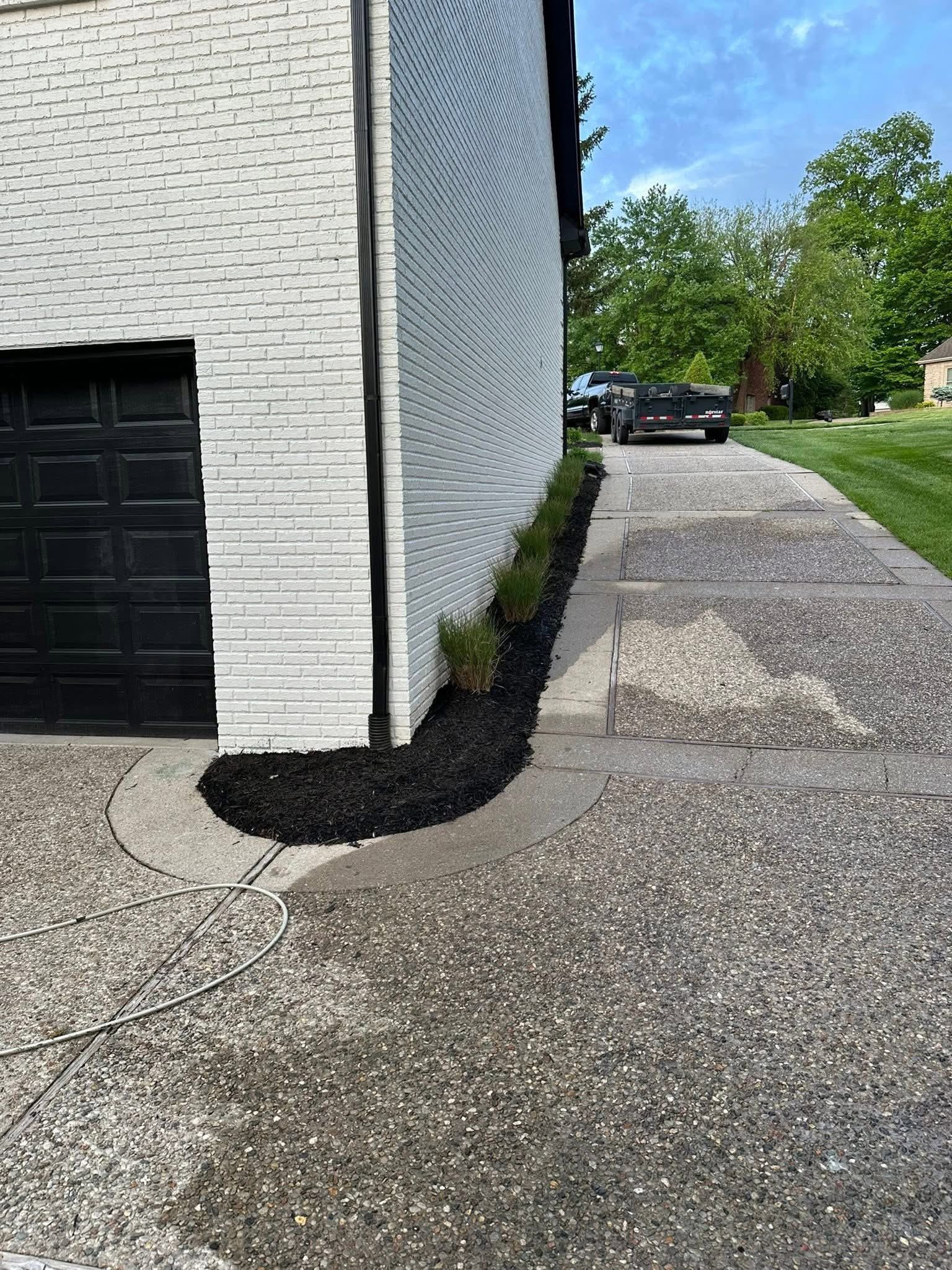 White brick building corner beside a paved driveway and black garage door, with a parked car in the distance.