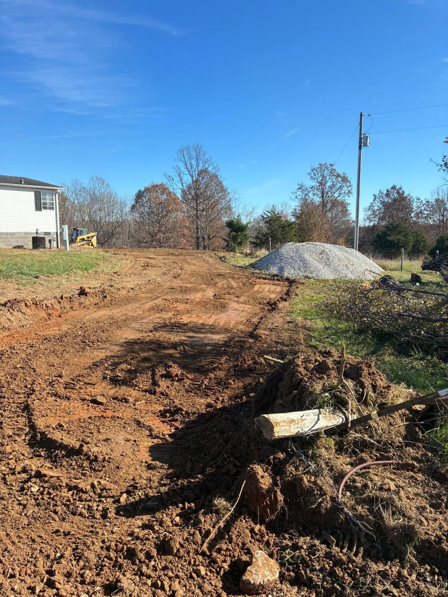Dirt construction site with a gravel pile, muddy ground, and a house at left under a clear blue sky