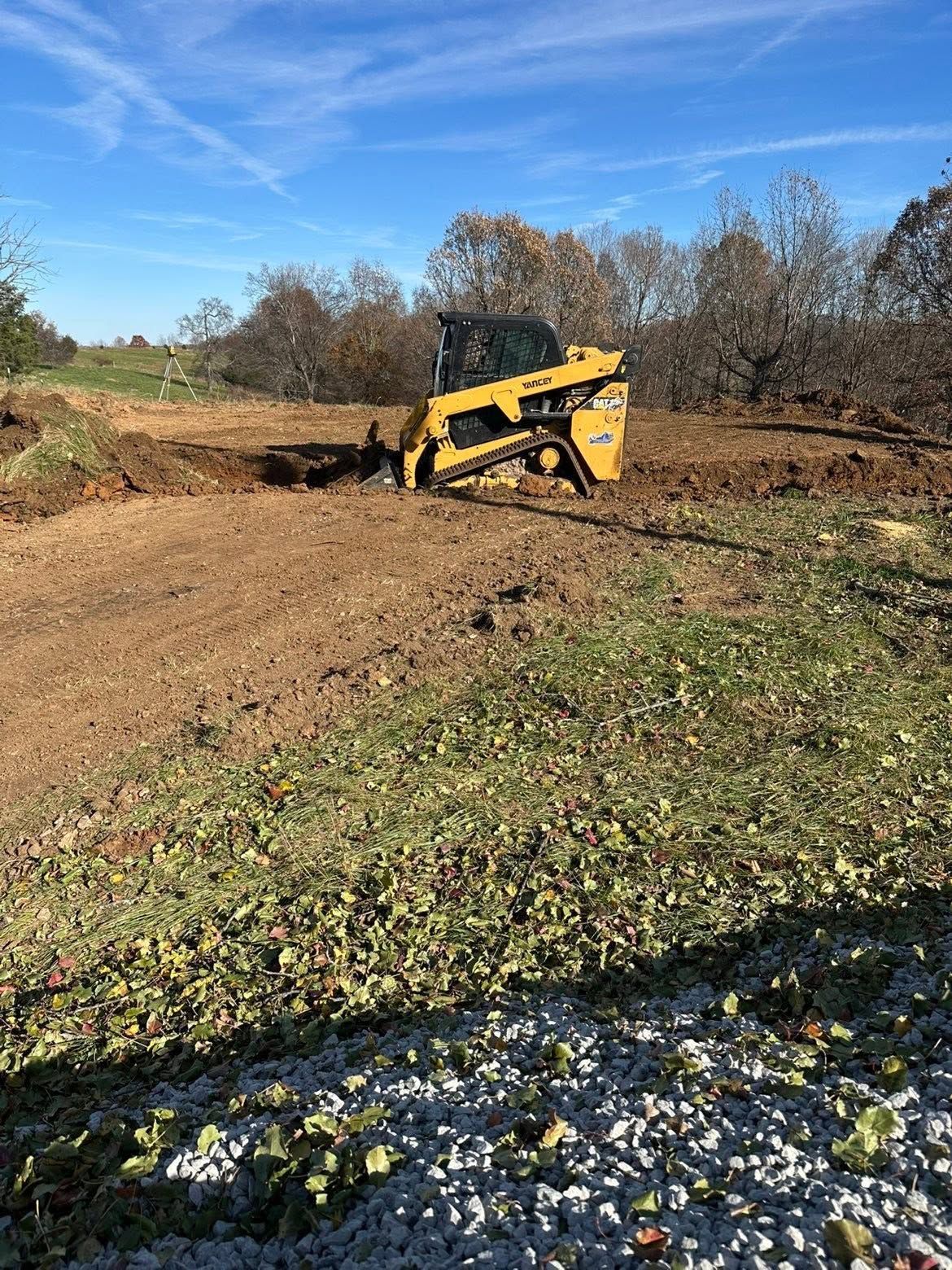 Yellow skid steer digging a dirt trench on a sunny rural lot
