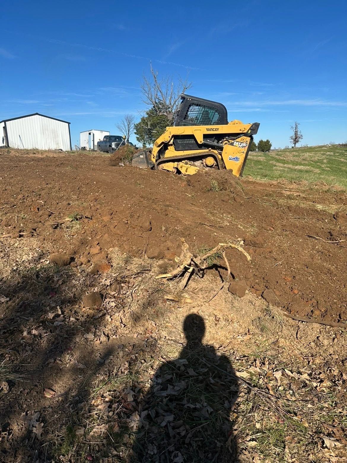 Yellow skid steer on a dirt hill near a white barn under a blue sky, with a person’s shadow in the foreground