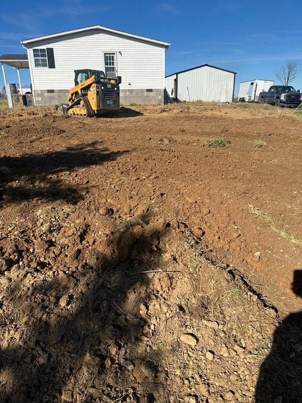 A small excavator sits on a dirt lot beside a white house and outbuilding under a clear blue sky.