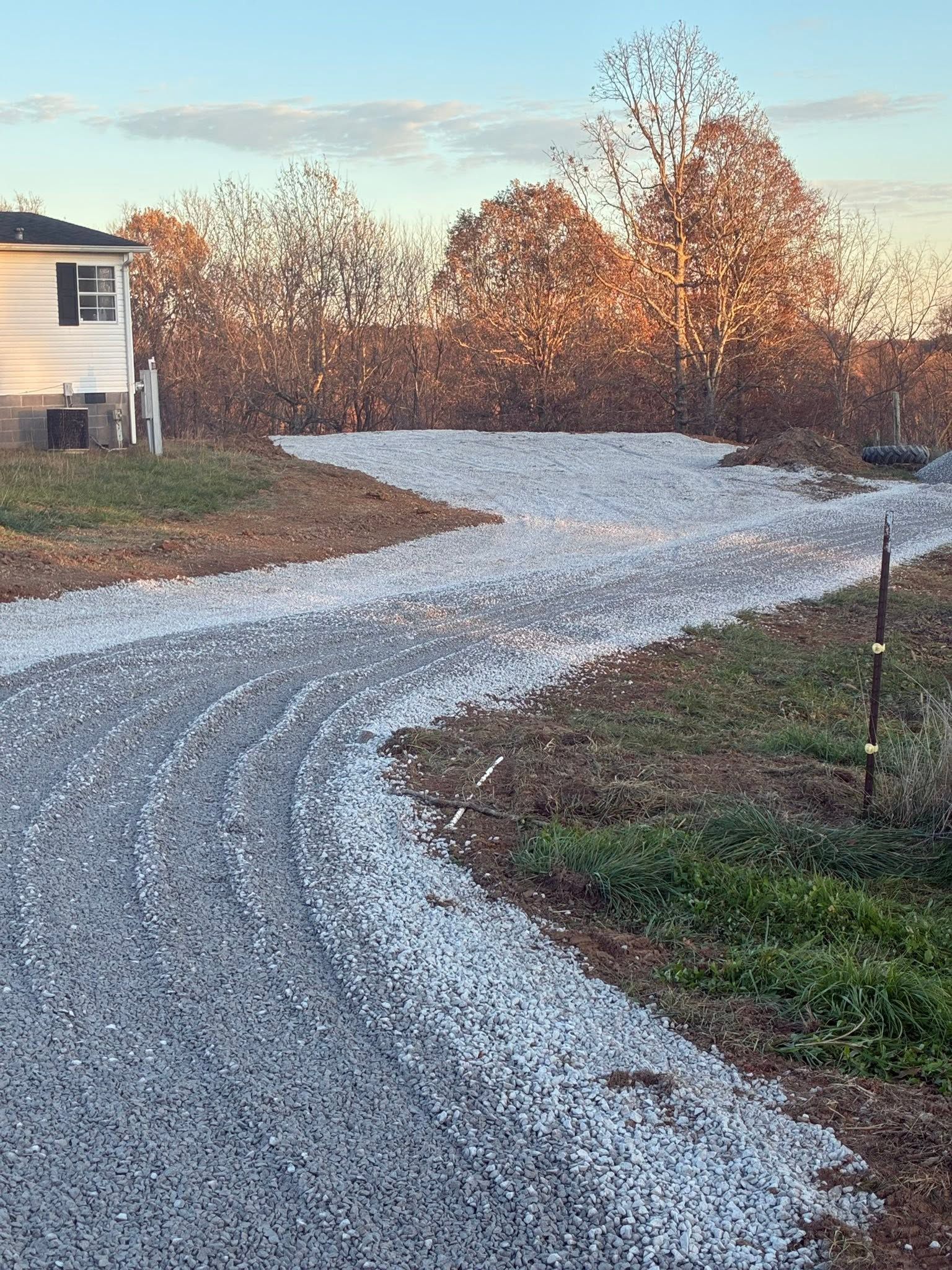 Gravel driveway curving past a house, bordered by autumn trees and brown grass.