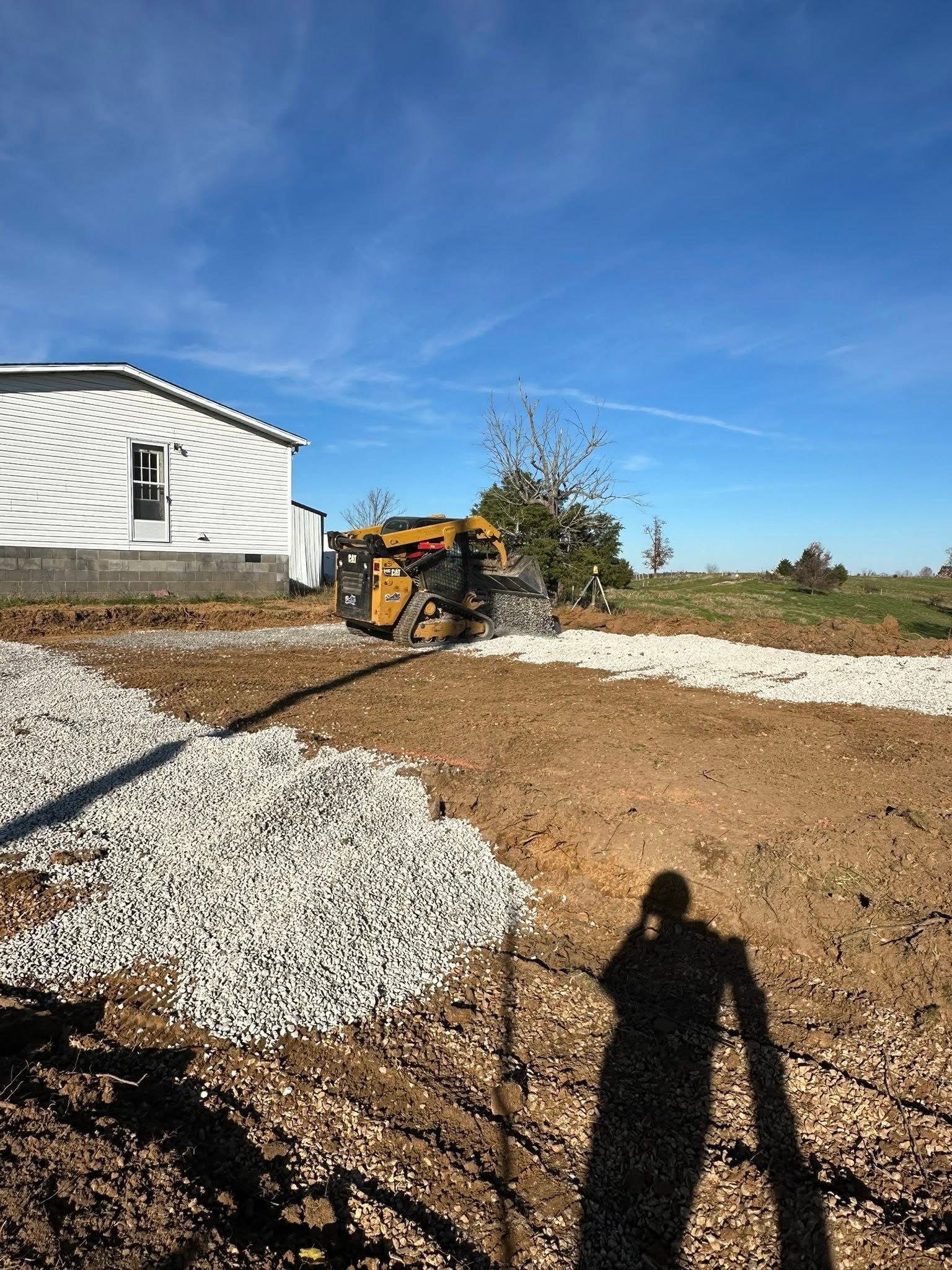 Construction equipment grading a gravel driveway beside a white house under a blue sky.